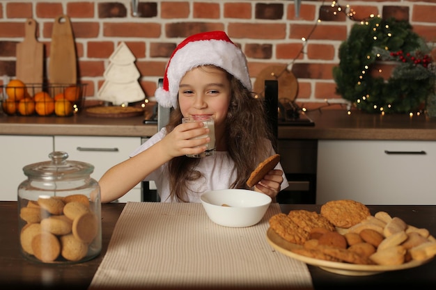 Ein Mädchen mit Weihnachtsmannmütze in der Küche hält Kekse und ein Glas Milch in den Händen