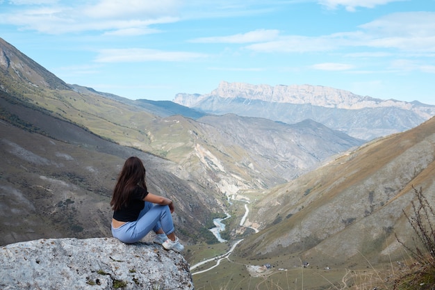 Ein Mädchen sitzt am Rand einer Klippe und schaut in die Ferne, in der Ferne ein Fluss und eine Schlucht, gute Laune, Wolken und eine Berglandschaft