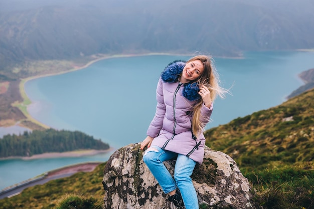 Ein Mädchen sitzt auf einem Berg, der Wind entwickelt ihr Haar Ein Landschaftsblick auf die Caldera Lagoa do Fogo unter einem nebligen Himmel mit großer Tiefe