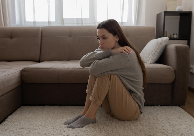 Ein Mädchen sitzt in Zivil auf einem weißen Teppich neben einem Sofa und umarmt ihre Knie