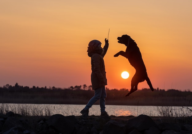 Ein Mädchen spielt mit einem Wachhund der Rasse Rottweiler vor dem Hintergrund eines Sees und Sonnenuntergangs