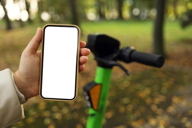 Ein Mädchen steht mit einem Roller und zeigt ein Telefon mit leerer weißer Beleuchtung in einem Herbstpark