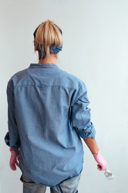 Ein Mädchen steht vor einer weißen Wand in einem blauen Hemd mit einem Pinsel in den Händen