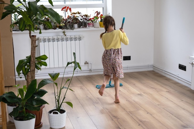 Ein Mädchen tanzt mit einem Wischmopp, um den Boden in einem neuen Haus zu reinigen, allgemeine Reinigung in einem leeren Raum, die Freude am Bewegen, Hilfe bei der Hausarbeit