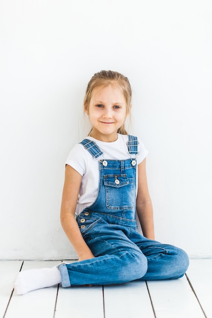 Ein Mädchen von europäischem Aussehen mit hell langen Haaren sitzt auf einem hellen Boden, gekleidet in ein T-Shirt und Jeans, Kindheit