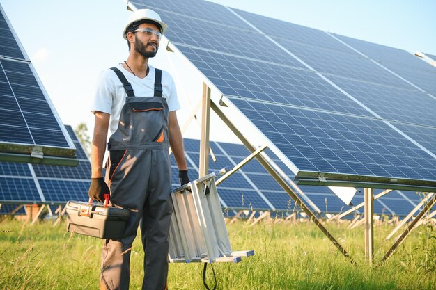 Ein männlicher arabischer Ingenieur in Helm und brauner Overall überprüft den Widerstand in Solarpanelen im Freien. Ein indischer Mann arbeitet an der Station.