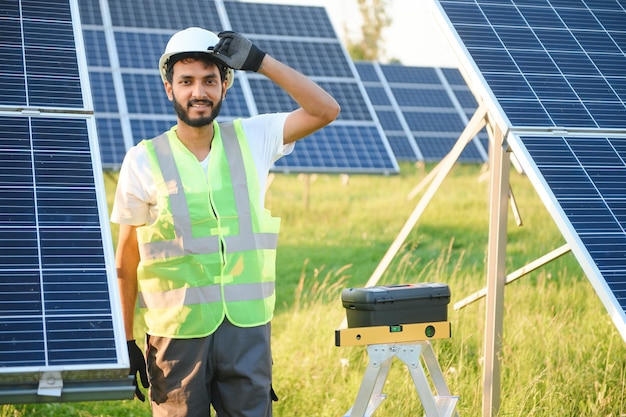 Ein männlicher arabischer Ingenieur in Helm und brauner Overall überprüft den Widerstand in Solarpanelen im Freien. Ein indischer Mann arbeitet an der Station.