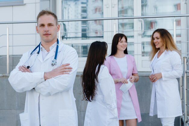 Ein männlicher Arzt blickt mit vor der Brust verschränkten Armen in die Kamera vor der Klinik. Im Hintergrund steht das Ärzteteam