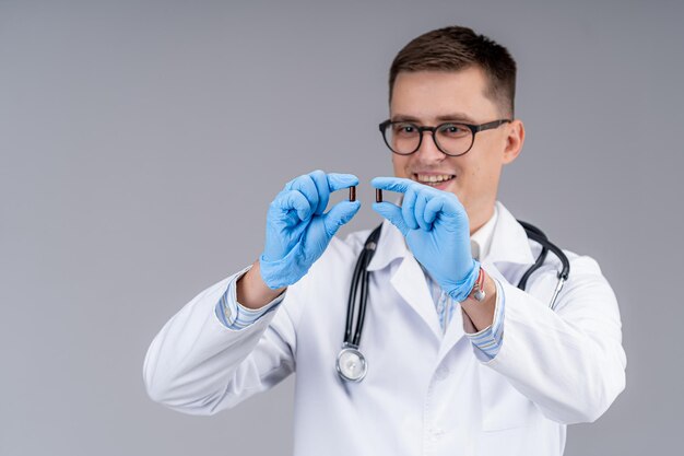 Ein männlicher Arzt im weißen Kittel vor einer grauen Wand. Sanitäter mit einem Stethoskop am Hals hält Pillen zwischen den Fingern.