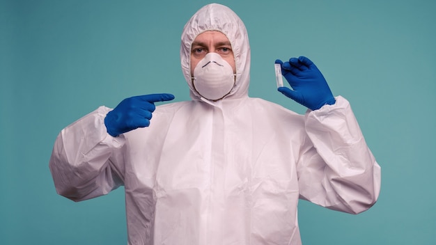 Ein männlicher Arzt in einem Schutzoverall und einer Maske, die mit einem Impfstoff in den Händen auf die Ampulle zeigt.