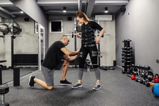 Ein männlicher Ausbilder hilft einer Frau im Stehen, einen Anzug für die EMS-Technologie in einem modernen Fitnessstudio-Konzept zu trainieren