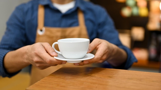 Ein männlicher Barista in Schürze, der eine Kaffeetasse hält oder einem Kunden im Café eine Tasse Cappuccino serviert