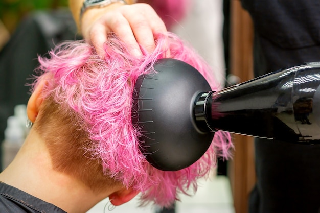 Ein männlicher Friseur, der in einem Schönheitssalon stilvolle rosa Haare der Kundin mit einem Fön trocknet