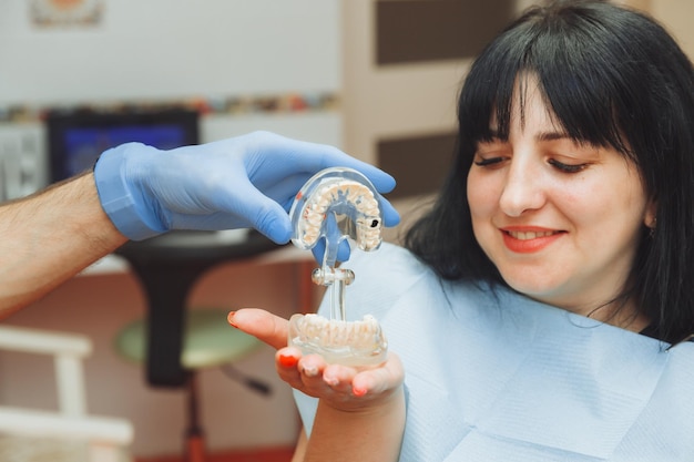 Ein männlicher kieferorthopädischer Zahnarzt zeigt einen künstlichen Kunststoffkiefer eines Patienten, der in einem Zahnarztstuhl sitzt, medizinische Behandlung von Zahnschmelz