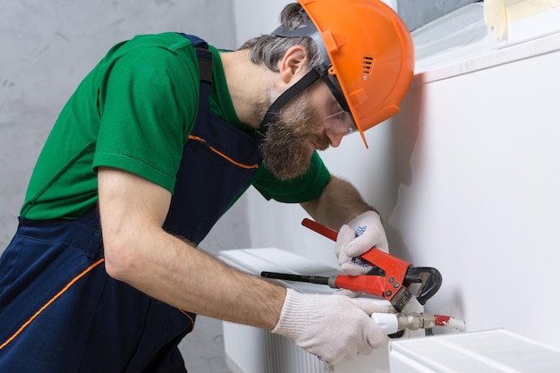 Ein männlicher Klempner installiert einen Heizkörper in der Heizungsanlage einer Wohnung Typ in Overall und Gasschlüssel