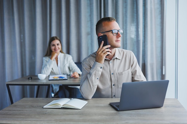 Ein männlicher Manager telefoniert mit einem Handy, während er an einem Tisch mit einem Computer und einem Notizblock sitzt. Arbeitsatmosphäre in einem Büro mit großen Fenstern