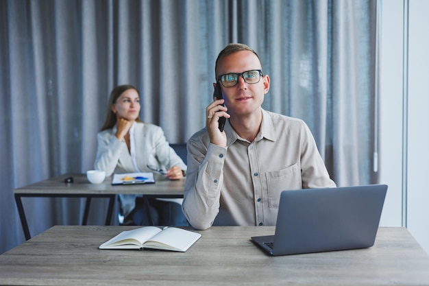 Ein männlicher manager telefoniert mit einem handy, während er an einem tisch mit einem computer und einem notizblock sitzt. arbeitsatmosphäre in einem büro mit großen fenstern
