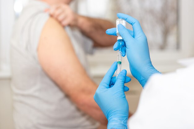 Ein männlicher Patient bereitet seine Schulter auf die Impfung vor. Nahaufnahme - Hände eines Arztes oder einer Krankenschwester in blauen Handschuhen mit einer medizinischen Spritze in einer Flasche Lösung