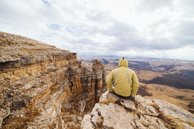 Ein männlicher Reisender in einer Jacke sitzt am Rand einer Klippe und genießt die Bergnatur und die saubere Luft