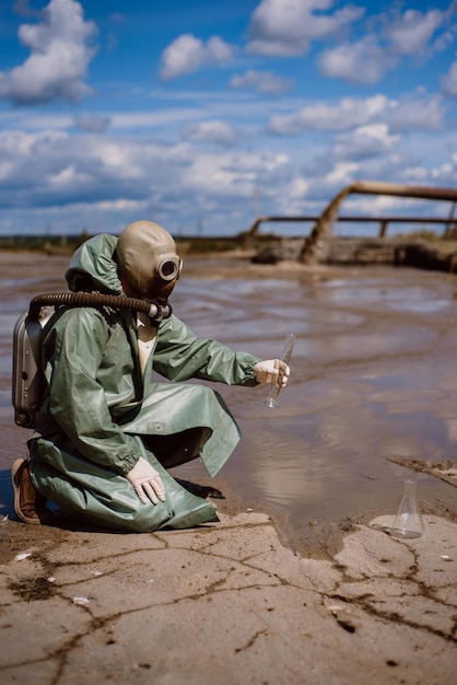 Ein männlicher Umweltschützer in einem grünen Schutzanzug und einer Gasmaske entnimmt eine Wasserprobe aus einem verschmutzten See für Forschungszwecke. Abfallproduktion. Wissenschaftler führt toxikologische Untersuchungen von Giftmüll durch