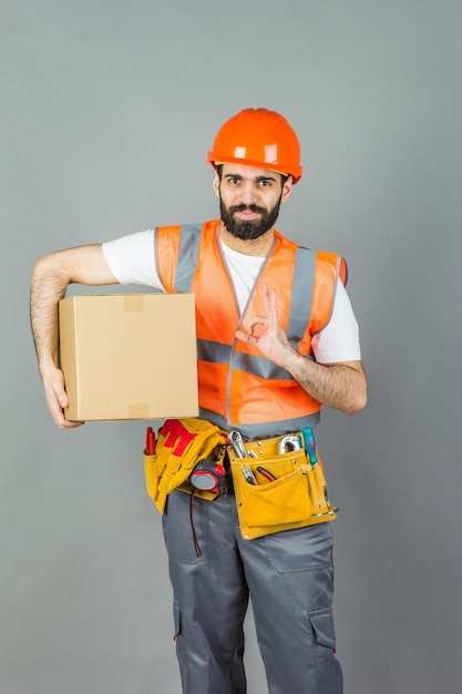 Ein Manbuilder in einem orangefarbenen Helm mit einem Karton in den Händen auf grauem Hintergrund