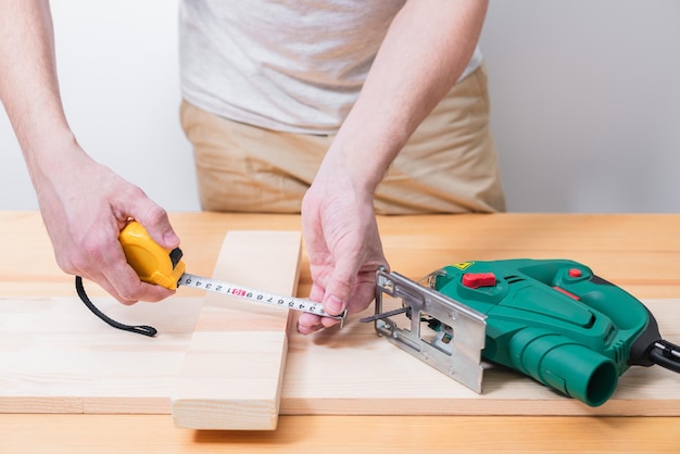 Ein Mann arbeitet mit einer elektrischen Stichsäge für Holz auf einem Holztisch mit und ohne Handschuhe und misst mit einem Maßband