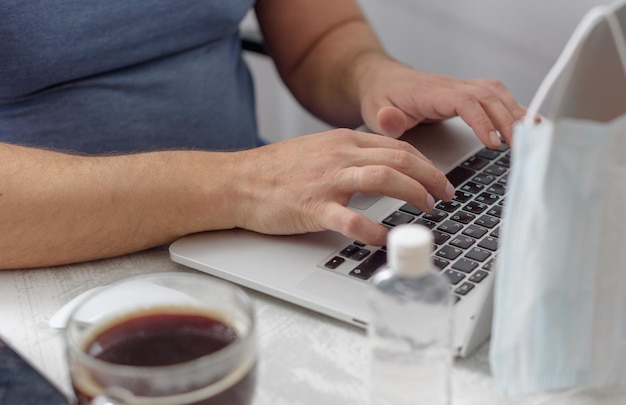 Ein mann arbeitet zu hause mit einem laptop, einem schützenden gesichtsmaskenkaffee und einer desinfektionsmittelquarantäne aufgrund der coronavirus-pandemie