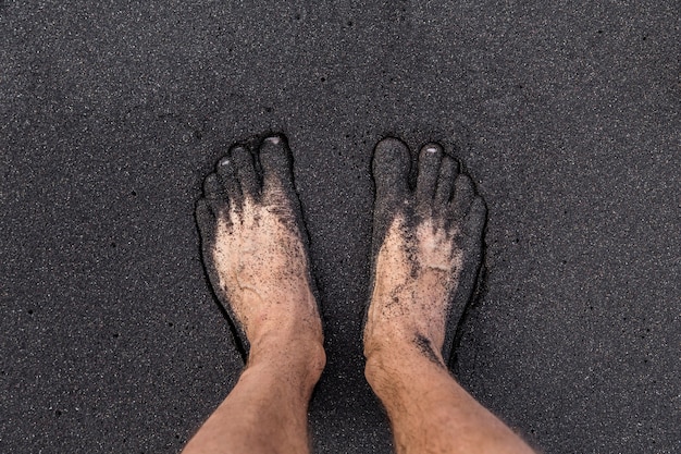 Ein Mann, der barfuß auf vulkanischem schwarzem Sand am Strand von Teneriffa steht. Zwei Fuß vom Sand bedeckt.