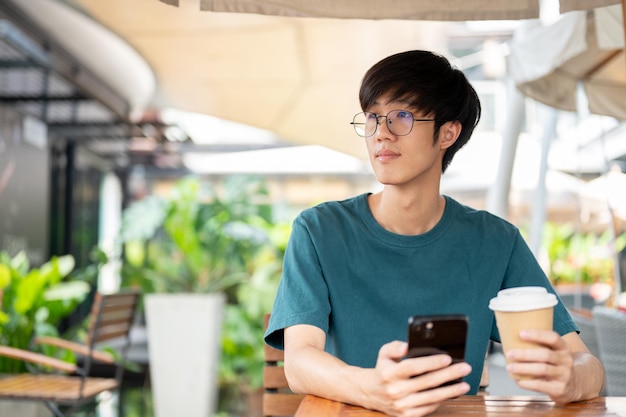 Ein Mann, der eine Kaffeetasse und ein Smartphone in der Hand hält und an einem Tisch im Freien eines Cafés wegblickt