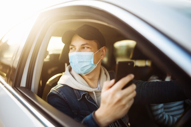 Ein Mann, der eine medizinische Maske trägt, hält ein Handy in der Hand, während er ein Auto fährt