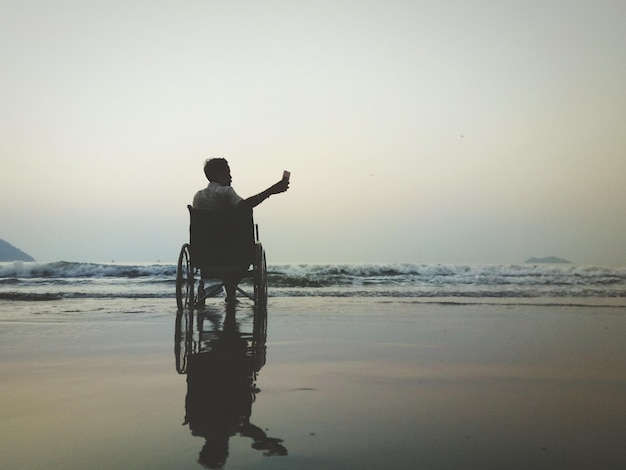 Ein Mann, der während des Sonnenuntergangs auf einem Rollstuhl am Strand sitzt und ein Selfie macht