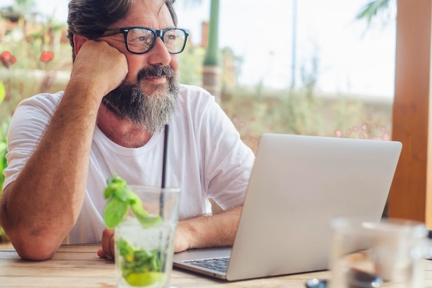 Ein Mann entspannt sich und träumt an der Bar mit Computer und Cocktail auf dem Tisch. Sommerurlaub, Urlaubsreisen und digitaler Nomade, Arbeiter, Job, Lebensstil, Person, heiterer Gesichtsausdruck, männliches Alter