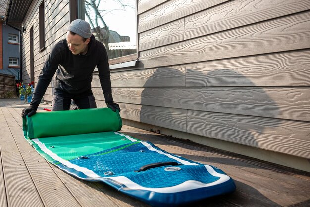 Ein Mann faltet ein aufblasbares Standup-Paddleboard SUP auf einem Holzdeck neben einem modernen Haus