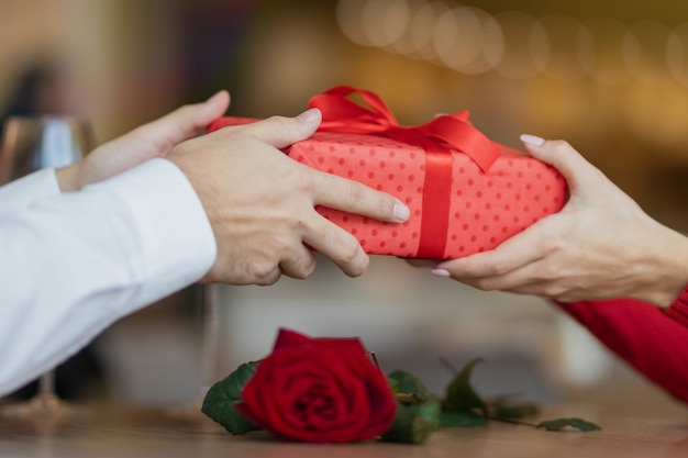 Ein Mann gibt seiner Freundin eine Geschenkbox mit einem roten Band. . Warmer und schöner Hintergrund eines Restaurants. Zwei Gläser Wein und eine Rose auf dem Tisch des Cafés. Valentinstag Konzept.