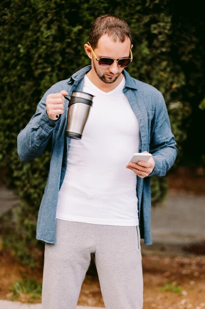 Foto ein mann hält eine kaffeetasse, während er einen handy benutzt und gegen bäume steht