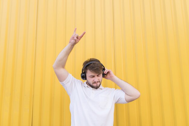 Ein Mann hört Rockmusik in Kopfhörern an einer gelben Wand und zeigt ein Heavy-Metal-Schild. Schwermetallsymbol