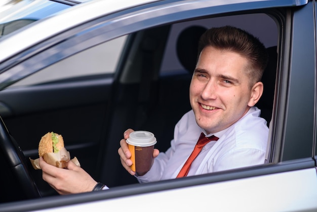 Ein Mann im Anzug trinkt Kaffee und isst einen Burger im Auto.