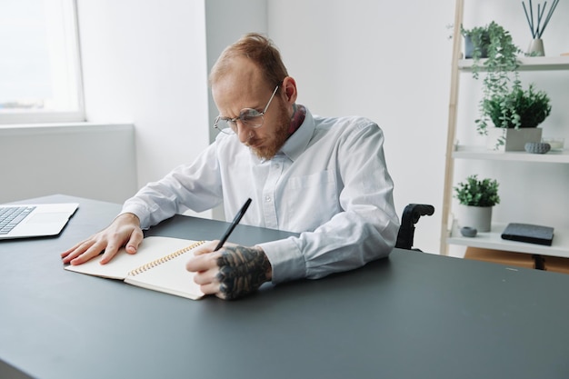 Ein Mann im Rollstuhl, ein Geschäftsmann im Büro, arbeitet an einem Laptop und schreibt einen Plan in ein Notizbuch. Nachdenklichkeit, Integration in die Gesellschaft, das Konzept der Arbeit einer Person mit Behinderungen