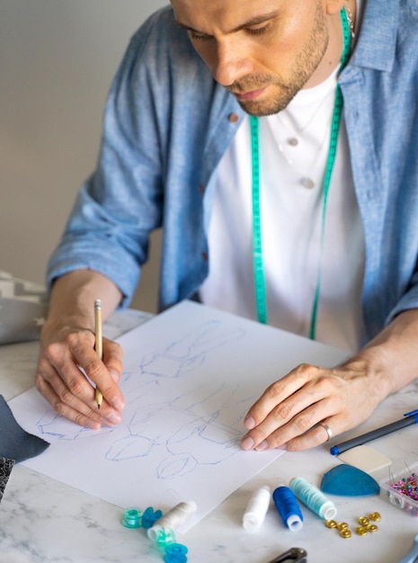 Ein Mann in einem blauen Hemd fertigt eine Skizze zum Nähen von Kleidung als Hobby an. Ein autodidaktischer Näher mit einem Maßband um den Hals arbeitet am Tisch. Designer zeichnet eine Skizze für ein neues Kostümprojekt