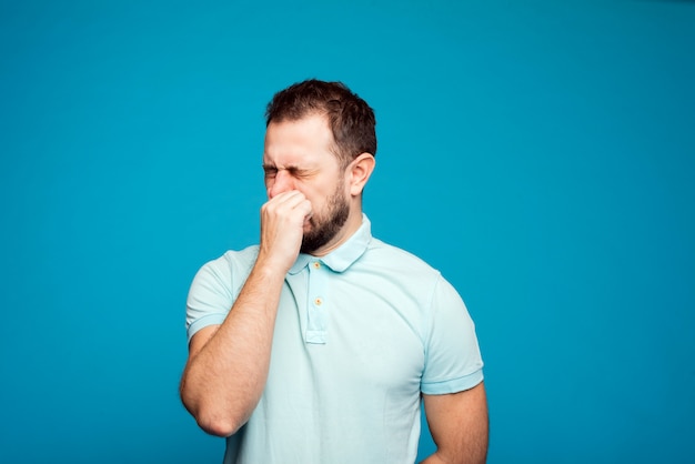 Ein Mann in einem blauen T-Shirt auf einem blauen Hintergrund niest in seiner Hand. Allergiekonzept. Nehmen Sie Allergiemedikamente