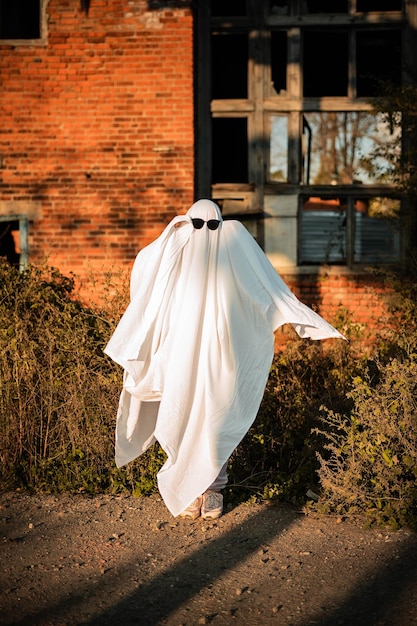 Foto ein mann in einem geisterkostüm aus bettlaken und sonnenbrille steht in der nähe eines verlassenen gebäudes ghost challenge 2021 spooky season halloween feiern