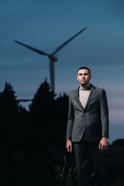 Ein Mann in einem grauen Anzug steht nach Sonnenuntergang neben einer Windmühle. Geschäftsmann in der Nähe von Windmühlen in der Nacht. Modernes Konzept der Zukunft.
