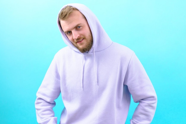 Ein Mann in einem lila Kapuzenpullover neigt den Kopf und blickt auf die Kamera auf blauem Hintergrund. Fröhlichkeit, die zuversichtlich, Ausdruck, freundlich, Freude, positiv, attraktiv, Lebensstil, modisch, lachen