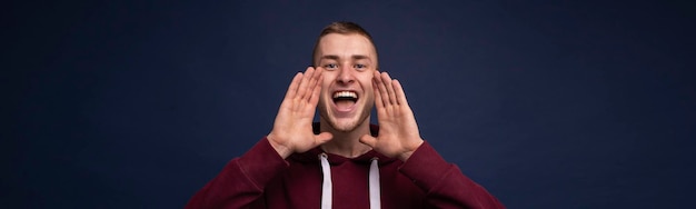 Ein Mann in einem roten Sweatshirt auf blauem Hintergrund schreit mit den Händen vor dem Gesicht