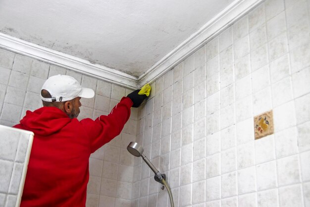 Foto ein mann in einem roten sweatshirt und einer weißen mütze reinigt schimmel aus einer badezimmerdecke