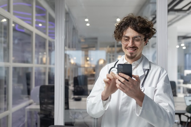 Ein Mann in einem weißen Labormantel und einem Stethoskop hält ein Handy in einer Arztpraxis einer Klinik