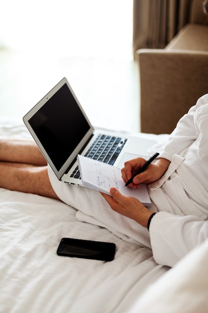 Ein Mann in einer Robe liegt mit einem Computer an den Füßen auf einem Bett. Freiberuflich, Arbeit von zu Hause aus. Kein Gesicht. Ein Mann in einem Hotel, der in einem Schlafzimmer an einem Computer arbeitet. Vertikal