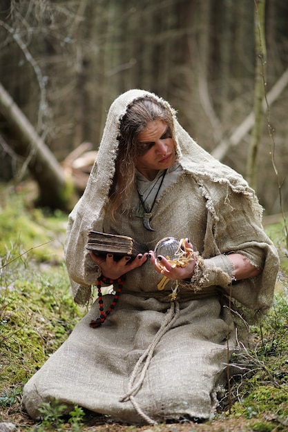 Ein Mann in einer Soutane verbringt ein Ritual in einem dunklen Wald mit einer Kristallkugel und einem Buch