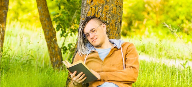 Ein Mann in Freizeitkleidung und Brille liest Literatur unter einem Baum Ein Hipster mit Dreadlocks