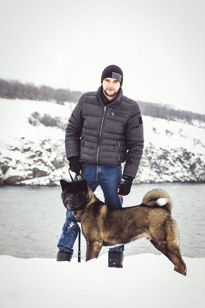 Ein Mann in Jacke und Strickmütze geht mit einem amerikanischen Akita-Hund durch einen verschneiten Wald